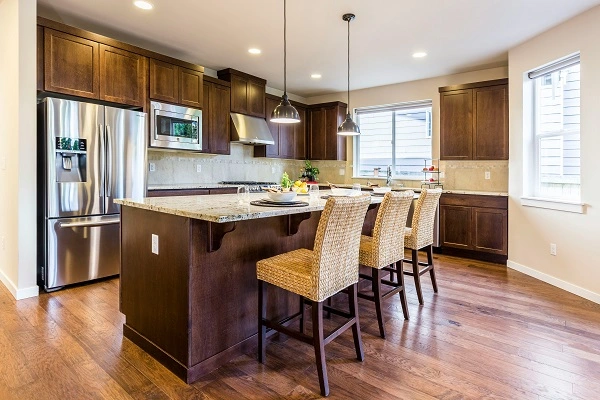 Cocina moderna fabricada en melamina en Style muebles Chile con gabinetes de madera oscura, isla con cubierta de granito beige y banquetas de ratán sobre piso de madera cálida